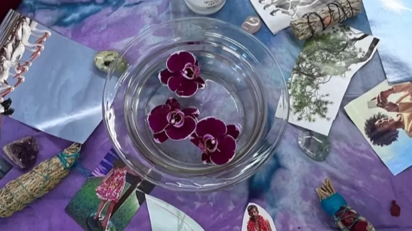 purple blanket with a bowl of water and red flowers floating, a candle, sage sticks and various pictures surrounding 